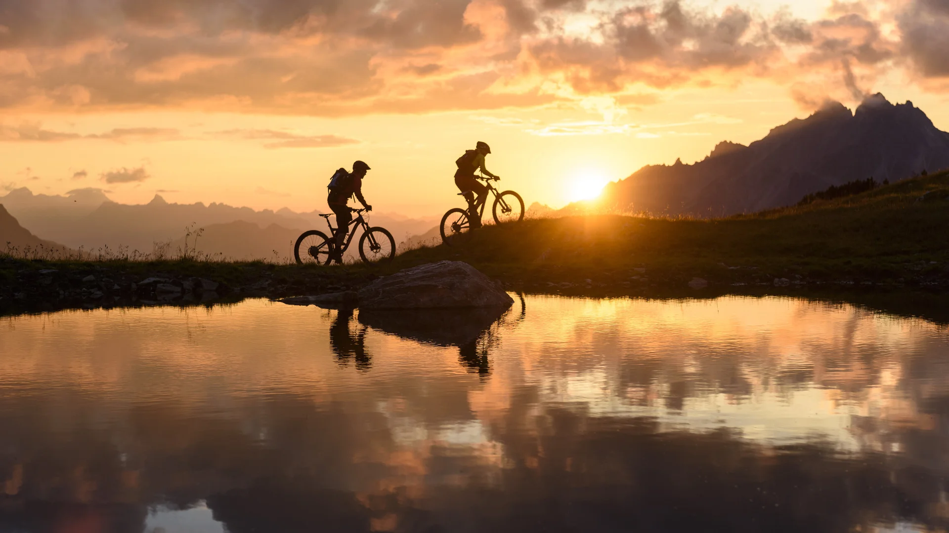 Zwei Personen fahren im Sonnenuntergang mit ihren Mountainbikes vor einer Bergkulisse. | © DAV/Wolfgang Ehn